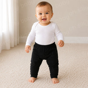 A baby standing while wearing Black Cotton Pants with a white long sleeve baby bodysuit
