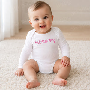 Baby girl wearing a white long sleeve Cleftstrong Heart baby bodysuit featuring a pink Cleftstrong heart design